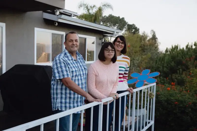 A family of three stands next to a white metal railing on the deck in the backyard of a house with trees around the exterior The family is smiling and looking towards the camera standing together